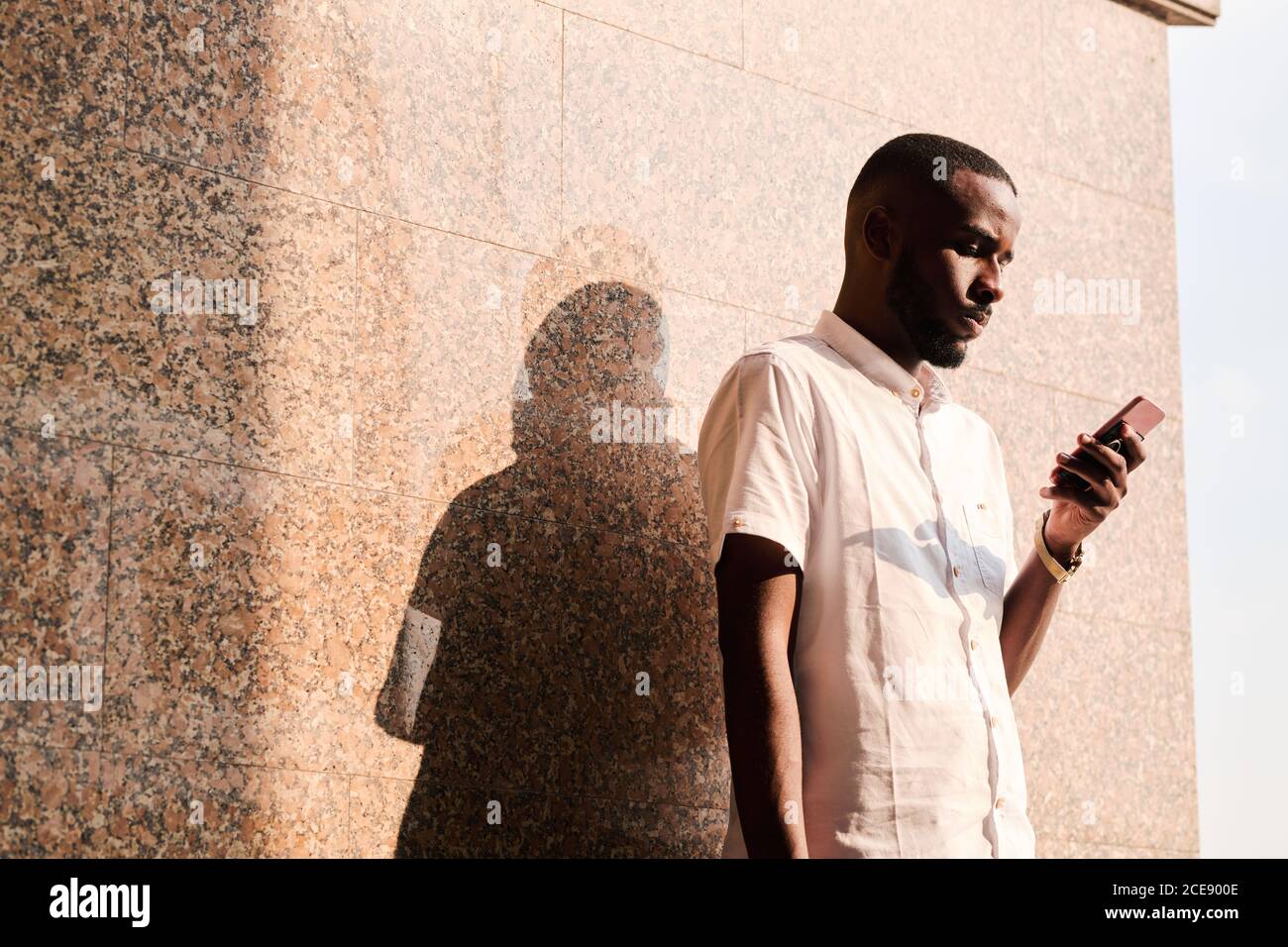 Concentrated young black man in white shirt standing at wall outdoors and reading sms on phone or checking mail Stock Photo
