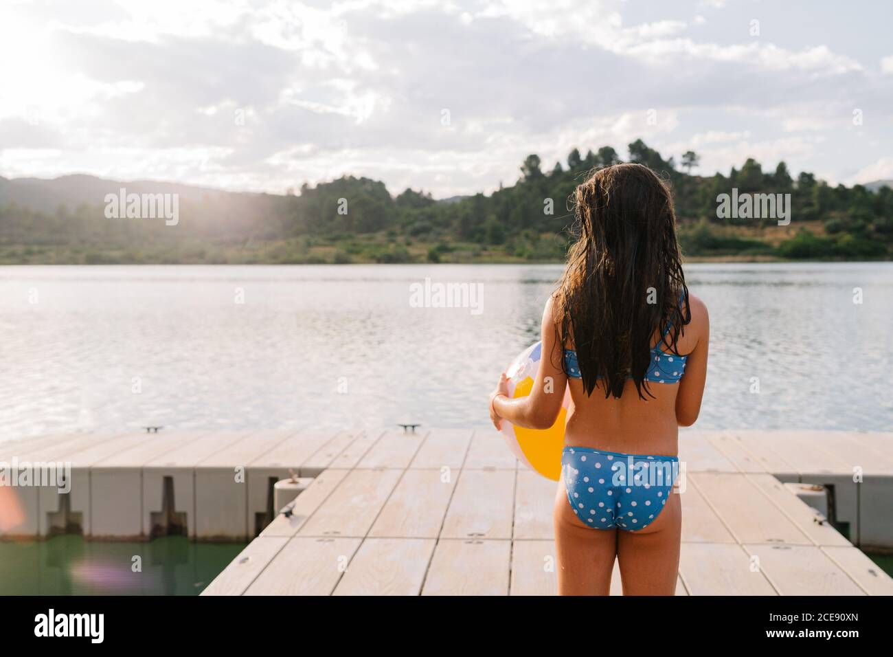 Back view of unrecognizable little girl in bikini playing with beach ball  standing in wooden pier near pond during summer holiday Stock Photo - Alamy