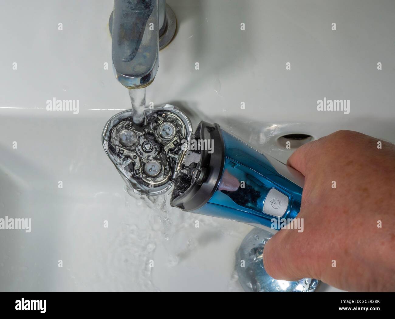 Closeup POV shot of a man’s hand holding a battery powered shaver under the flow of water from a bathroom sink tap, to wash away the dust and hair. Stock Photo