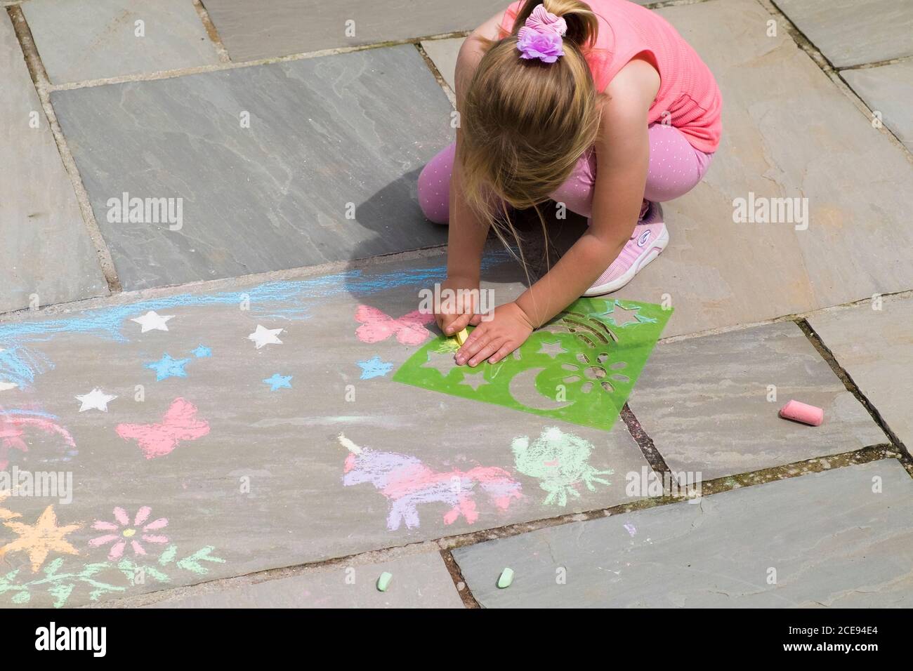 A five year old girl amusing herself busily using plastic stencils to chalk shapes on a flagstone floor. Stock Photo