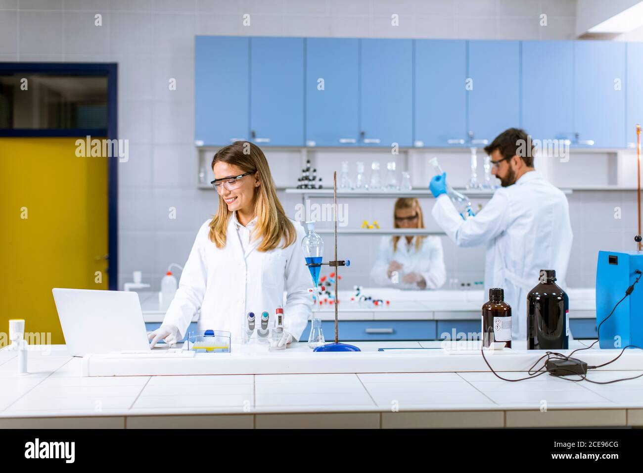 Cute female researcher in white lab coat and protective mask using laptop while working in the laboratory Stock Photo