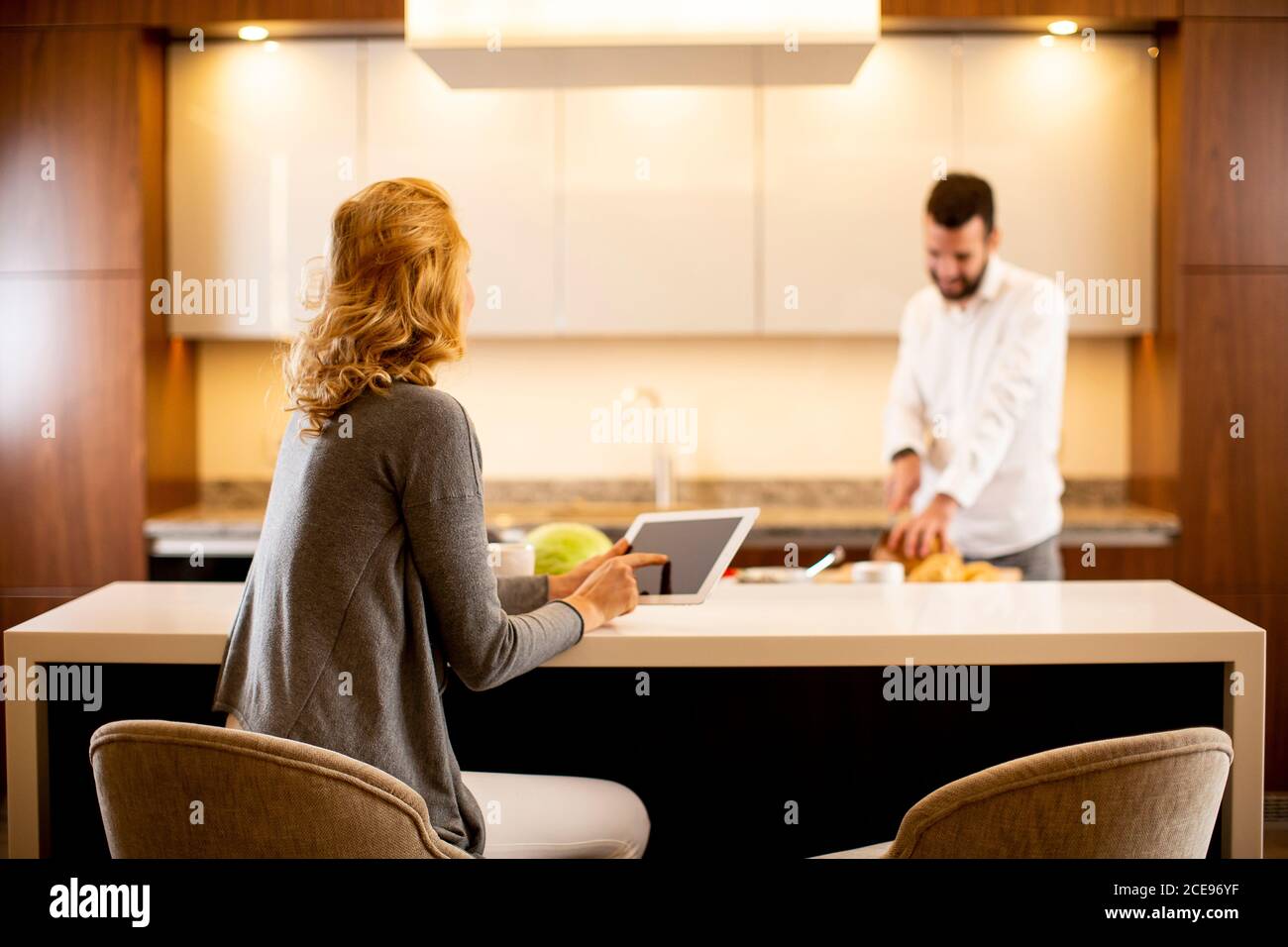 Young woman using tablet at the kitchen table while man preparing food Stock Photo