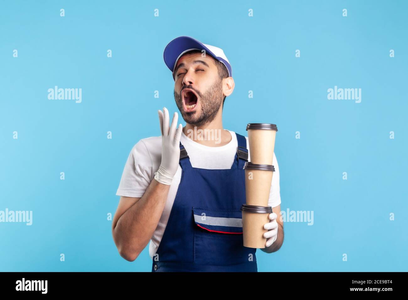 Delivery service. Tired sleepy courier man in blue overalls holding three coffee and yawning with exhausted bored expression, bringing drinks in dispo Stock Photo
