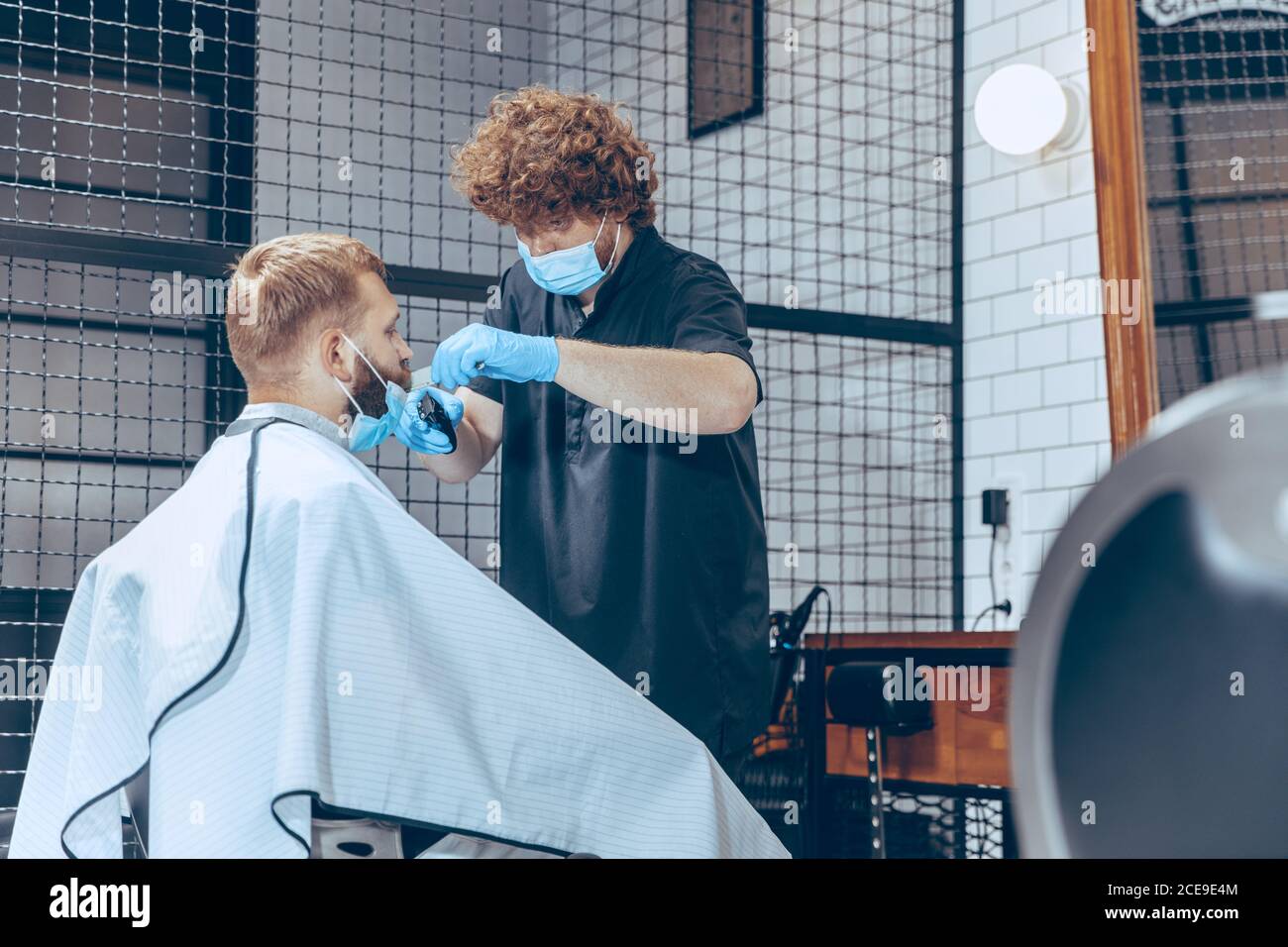 Man getting hair cut at the barbershop wearing mask during coronavirus pandemic. Professional barber wearing gloves. Covid-19, beauty, selfcare, style, healthcare and medicine concept. Stock Photo