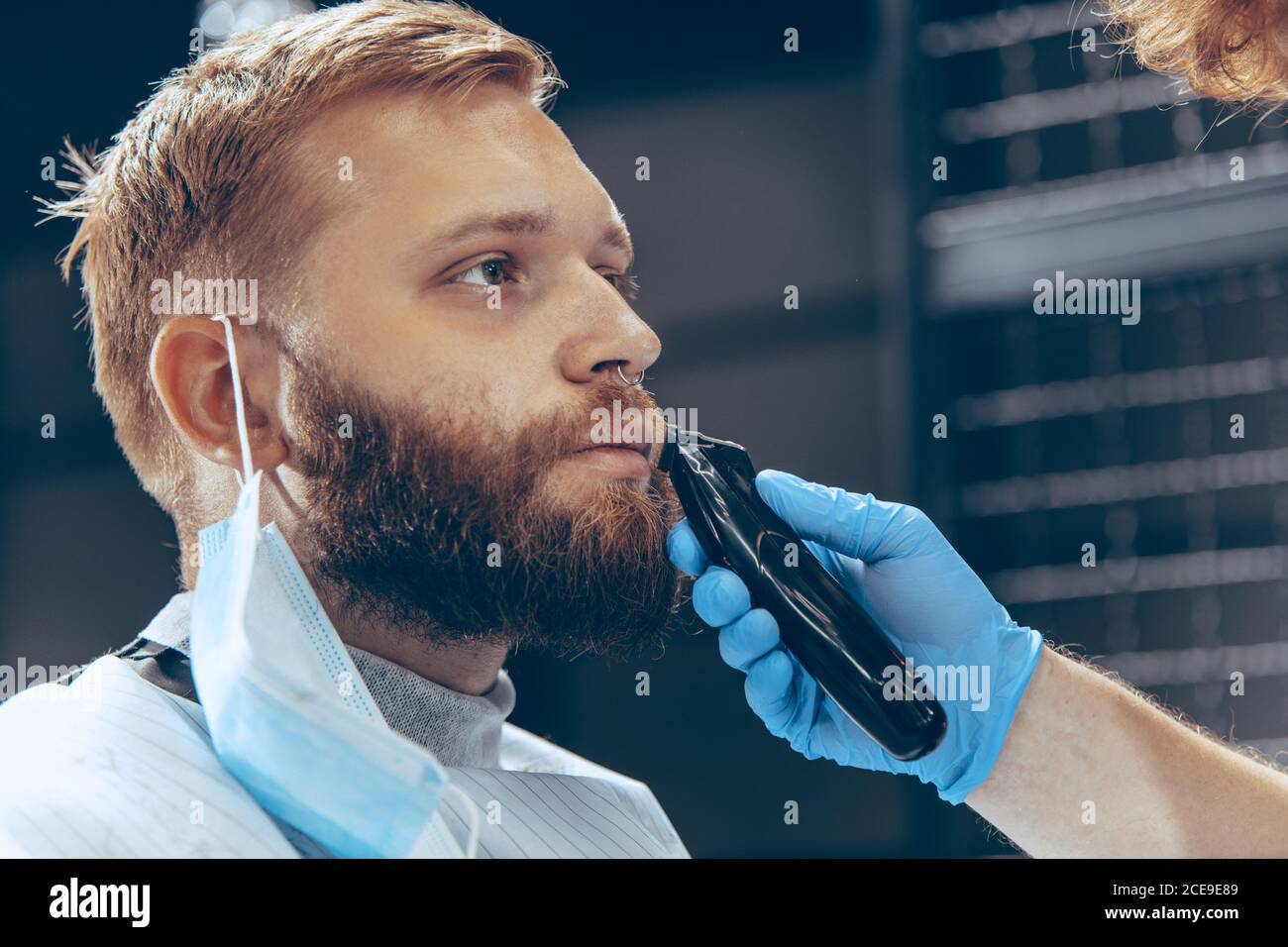 Close up man getting hair cut at the barbershop wearing mask during coronavirus pandemic. Professional barber wearing gloves. Covid-19, beauty, selfcare, style, healthcare and medicine concept. Stock Photo