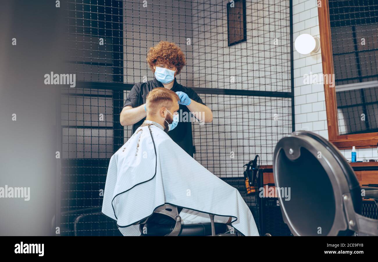 Man getting hair cut at the barbershop wearing mask during coronavirus pandemic. Professional barber wearing gloves. Covid-19, beauty, selfcare, style, healthcare and medicine concept. Stock Photo