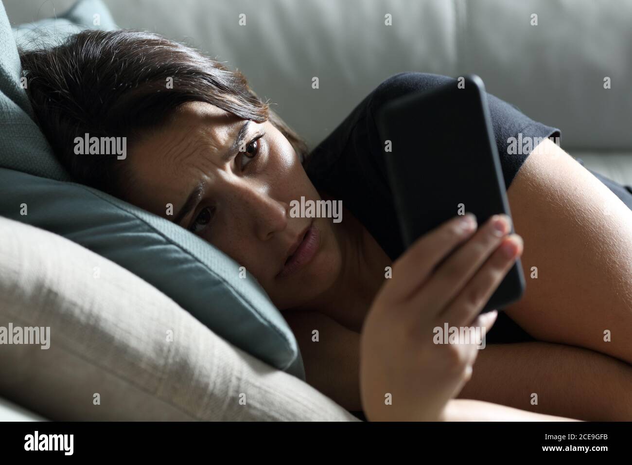 Sad woman checking phone in the dark lying on a sofa in the living room at home Stock Photo