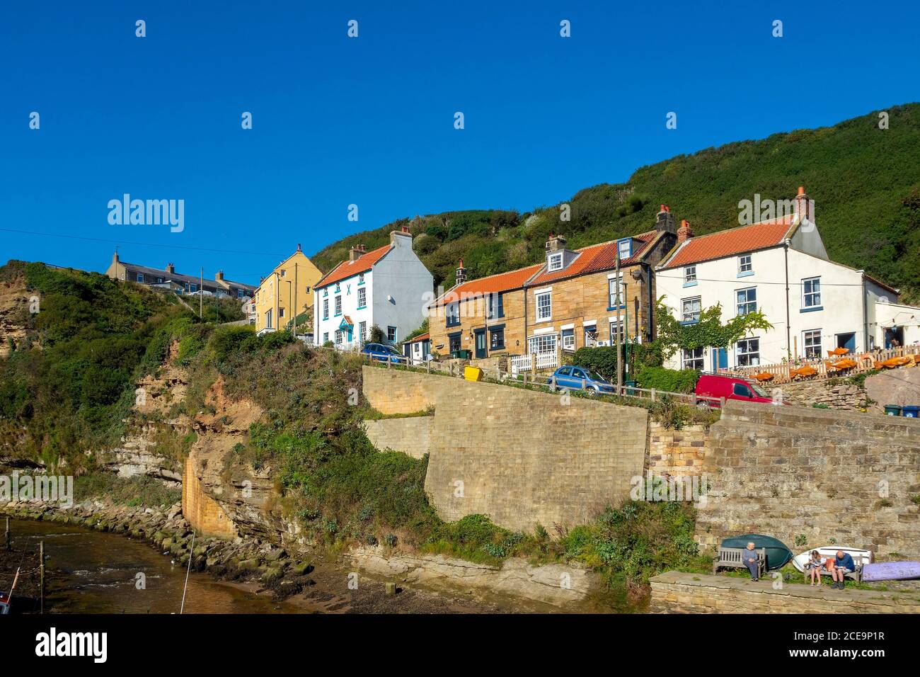 People sitting in the summer sunshine below the houses on the steep hill at Cowbar, Staithes on the North Sea Coastal path and Cleveland Way l Stock Photo