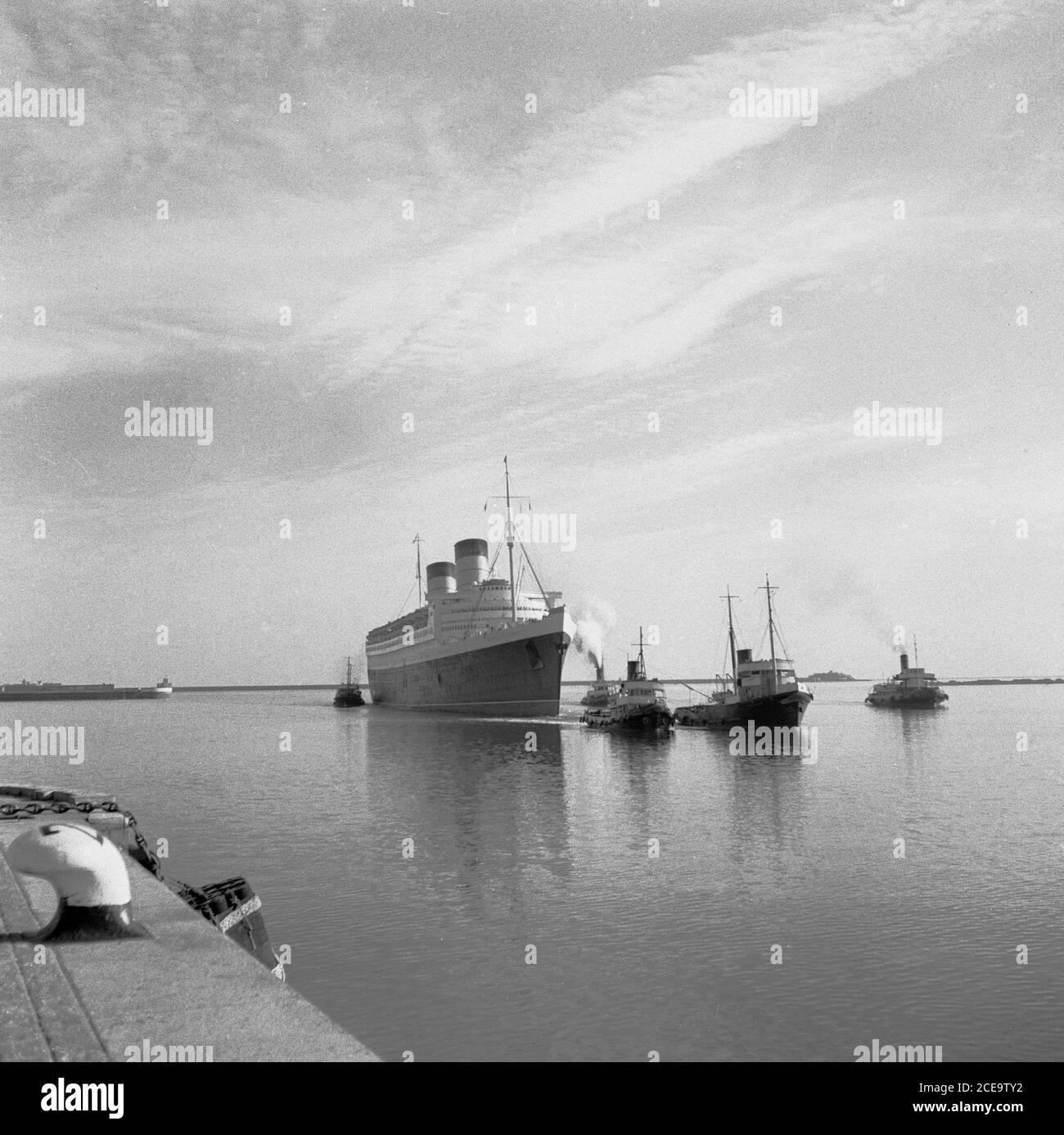 1950s, historical, tugboats pull the RMS Queen Elizabeth ocean liner into the calm waters of the French port of Cherbourg. Named after the British Queen Mother, it was the largest passenger liner ever built at that time (1938) and remained so for 56 years thereafter. She was used as a wartime troopship during WW2 and only became an ocean liner, as intended, in 1946. From then on, this luxury liner provided a weekly service between Southampton, UK and New York city in the USA, via Cherbourg, France.  In 1972  in Victoria Harbour, Hong Kong, while undergoing a refit, she caught fire and sank. Stock Photo