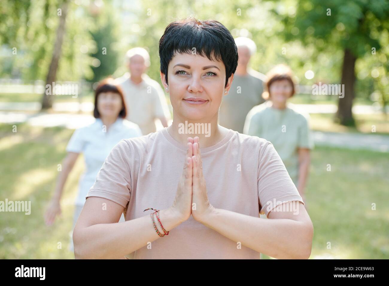 Portrait of content serene mature yoga instructor making Namaste gesture while standing in front of her students in park Stock Photo