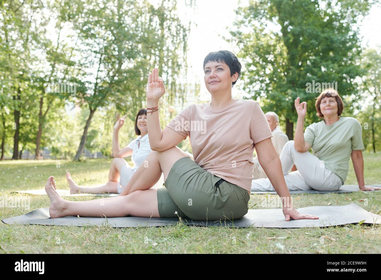 Attractive yoga coach with short hair sitting on mat and practicing marichis pose with students at group class Stock Photo