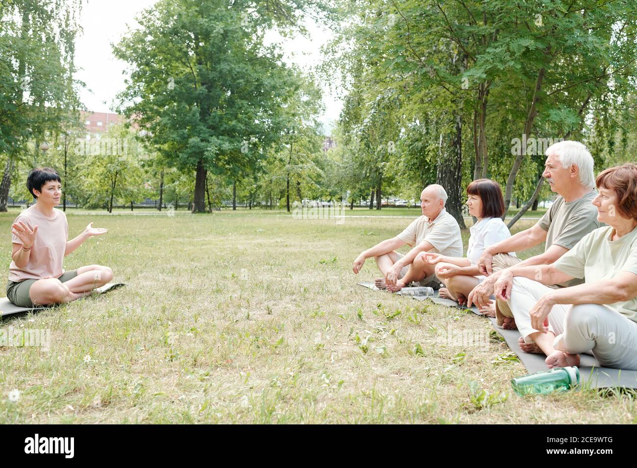 Short-haired yoga teacher sitting in lotus position and gesturing hands while talking to yoga students before class Stock Photo