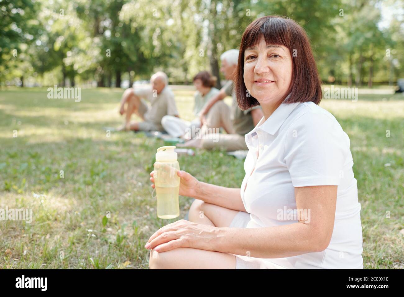 Portrait of content mature lady in white tshirt sitting on grass at outdoor training and drinking water Stock Photo