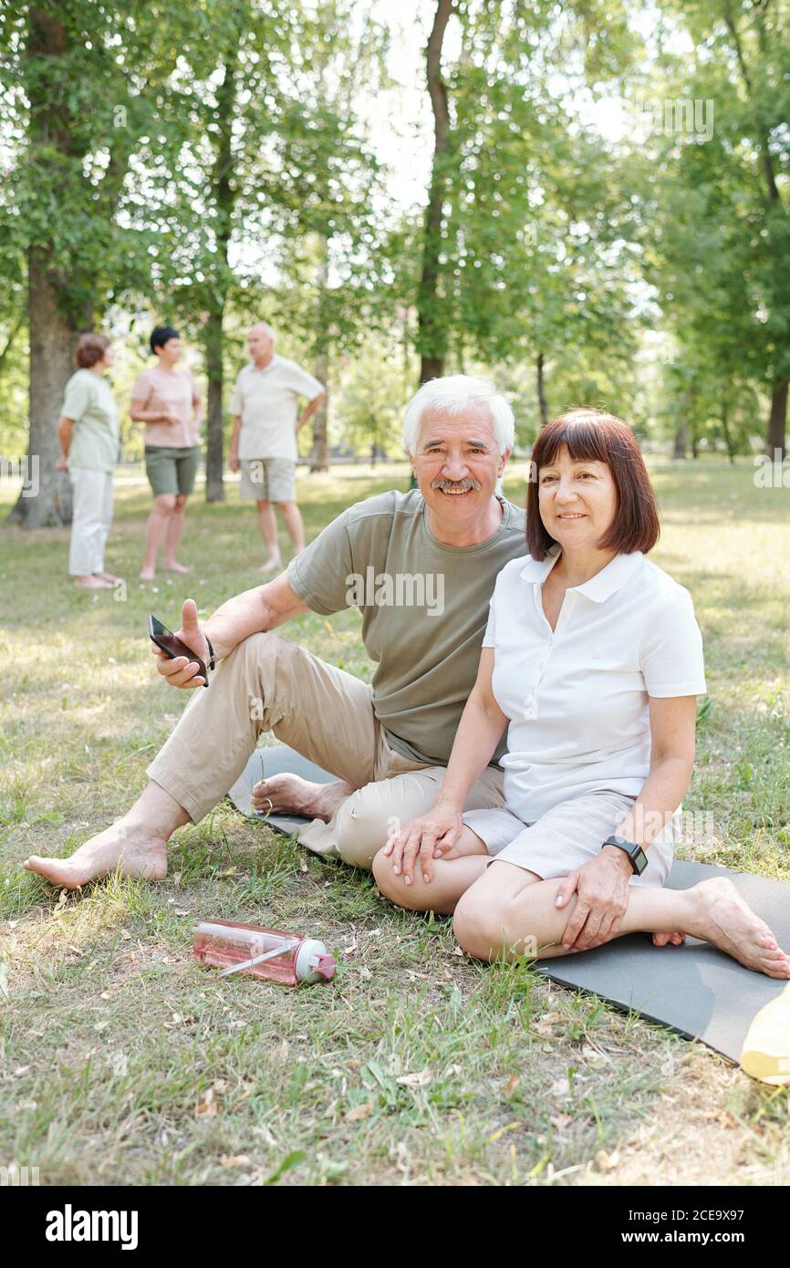 Portrait of positive active senior couple sitting with smartphone on yoga mat in park Stock Photo