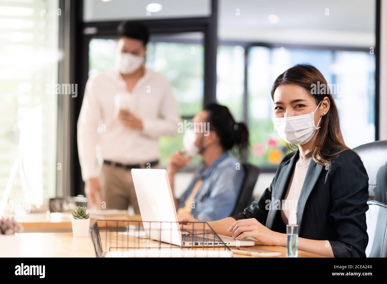 Portrait of asian office employee businesswoman wear protective face mask work in new normal office with interracial colleague in background as social Stock Photo