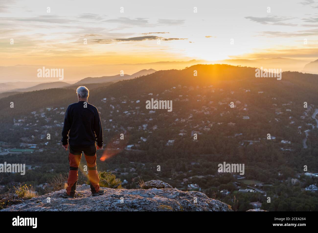 Back view of elderly man in casual outfit sitting on rocky cliff and admiring view of majestic mountains and beautiful evening sky Stock Photo