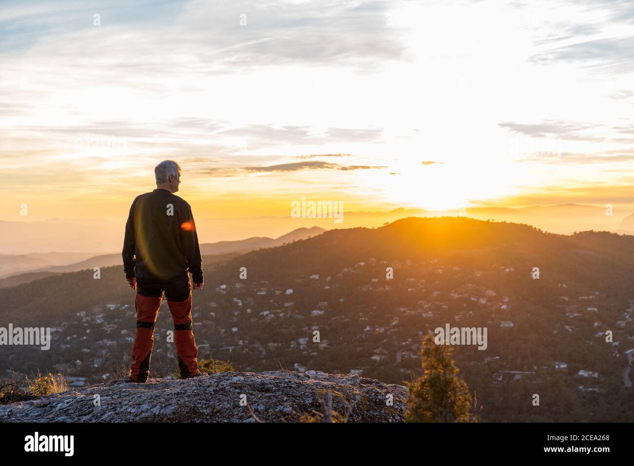 Back view of elderly man in casual outfit sitting on rocky cliff and admiring view of majestic mountains and beautiful evening sky Stock Photo