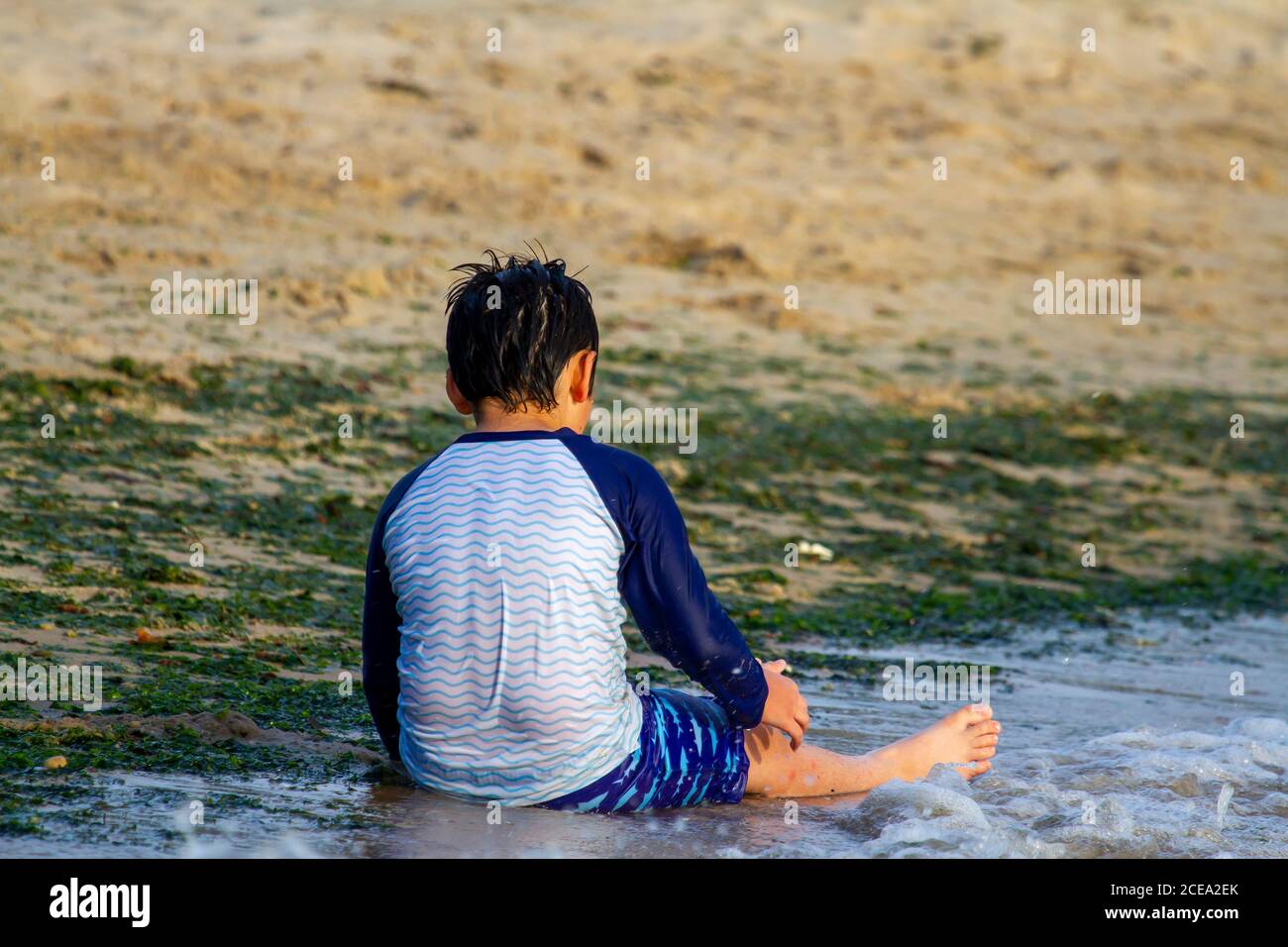 A young caucasian boy wearing rash guard long sleeve swimsuit is sitting on  sand by the sea. He is playing alone with the sand and water. Image was ta  Stock Photo 