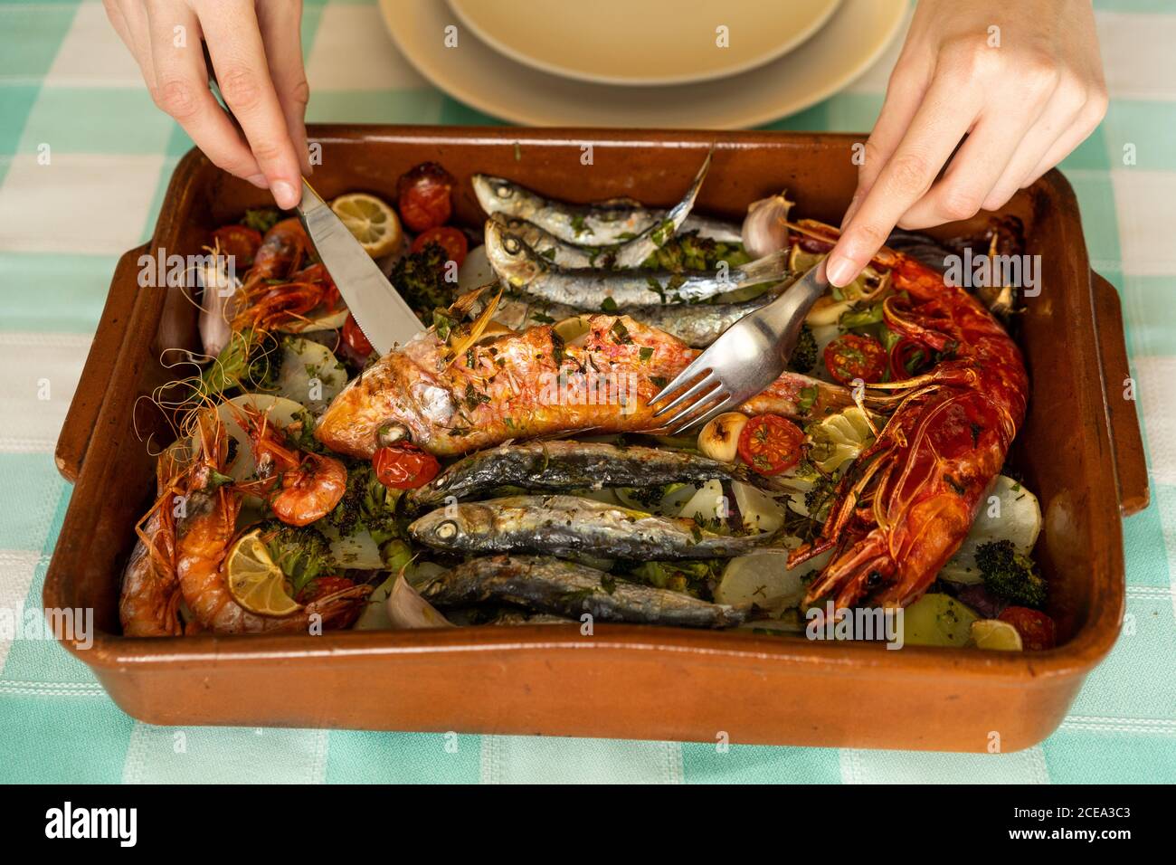 Hands of anonymous female using fork and knife to take delectable seafood dish from ceramic pan on table Stock Photo