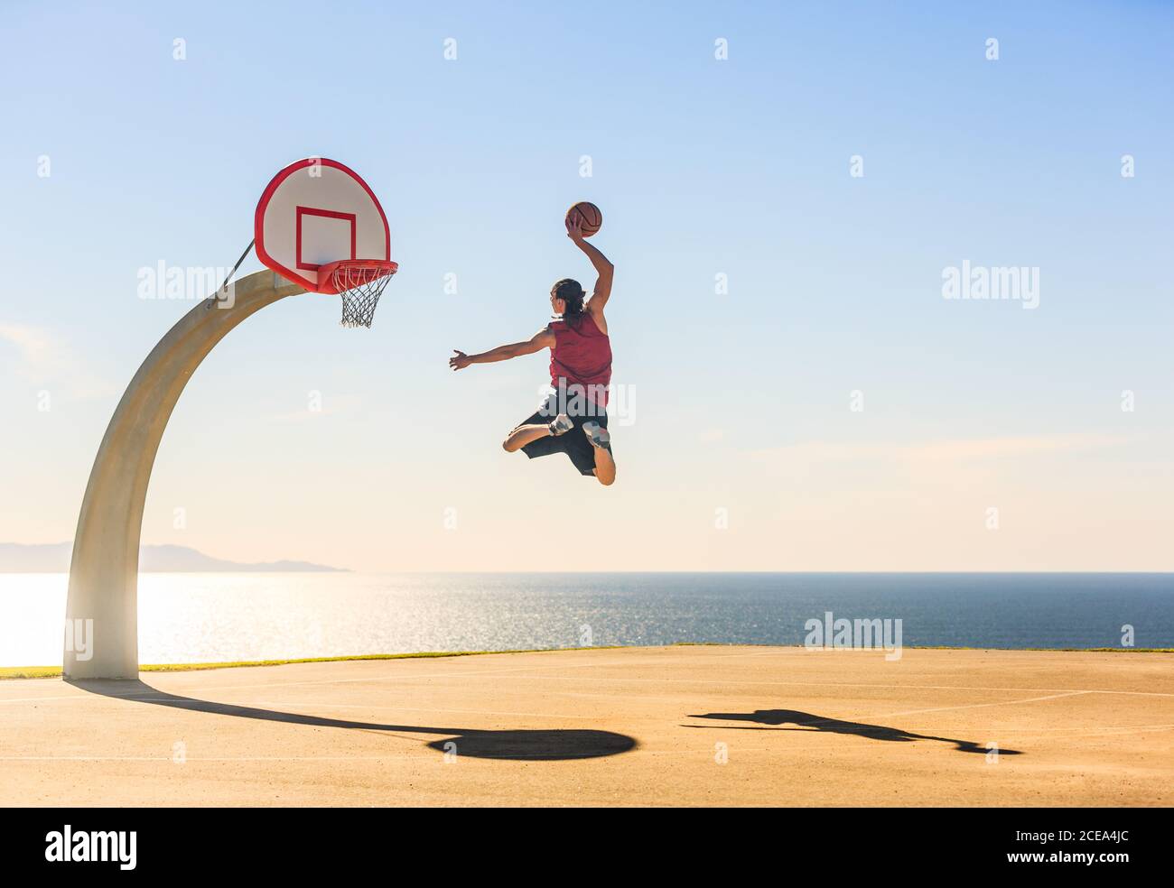 Basketball player scoring an amazing energetic dunk elevating at the street ball court with an ocean view as background. Sports, motivation and boost. Stock Photo