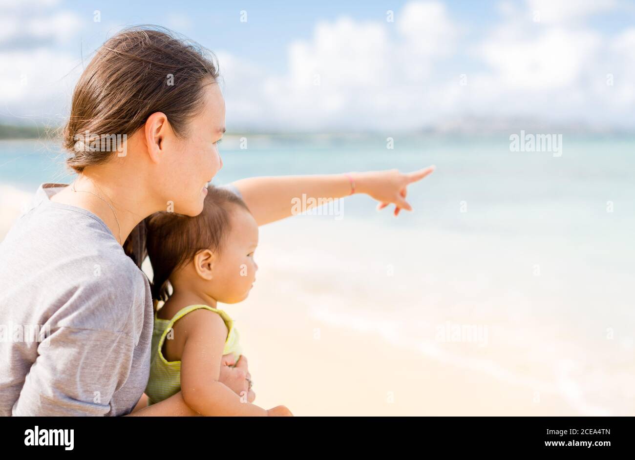 Mother and daughter family bonding on beautiful tropical beach during summer holiday vacation. Parent pointing out to the view showing her baby girl. Stock Photo