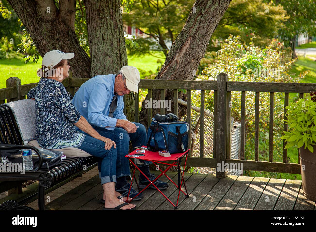 Chesapeake City, MD, USA 08/25/2020: An elderly caucasian couple wearing baseball hats shirts and jeans is sitting on pillows on a metal park bench in Stock Photo