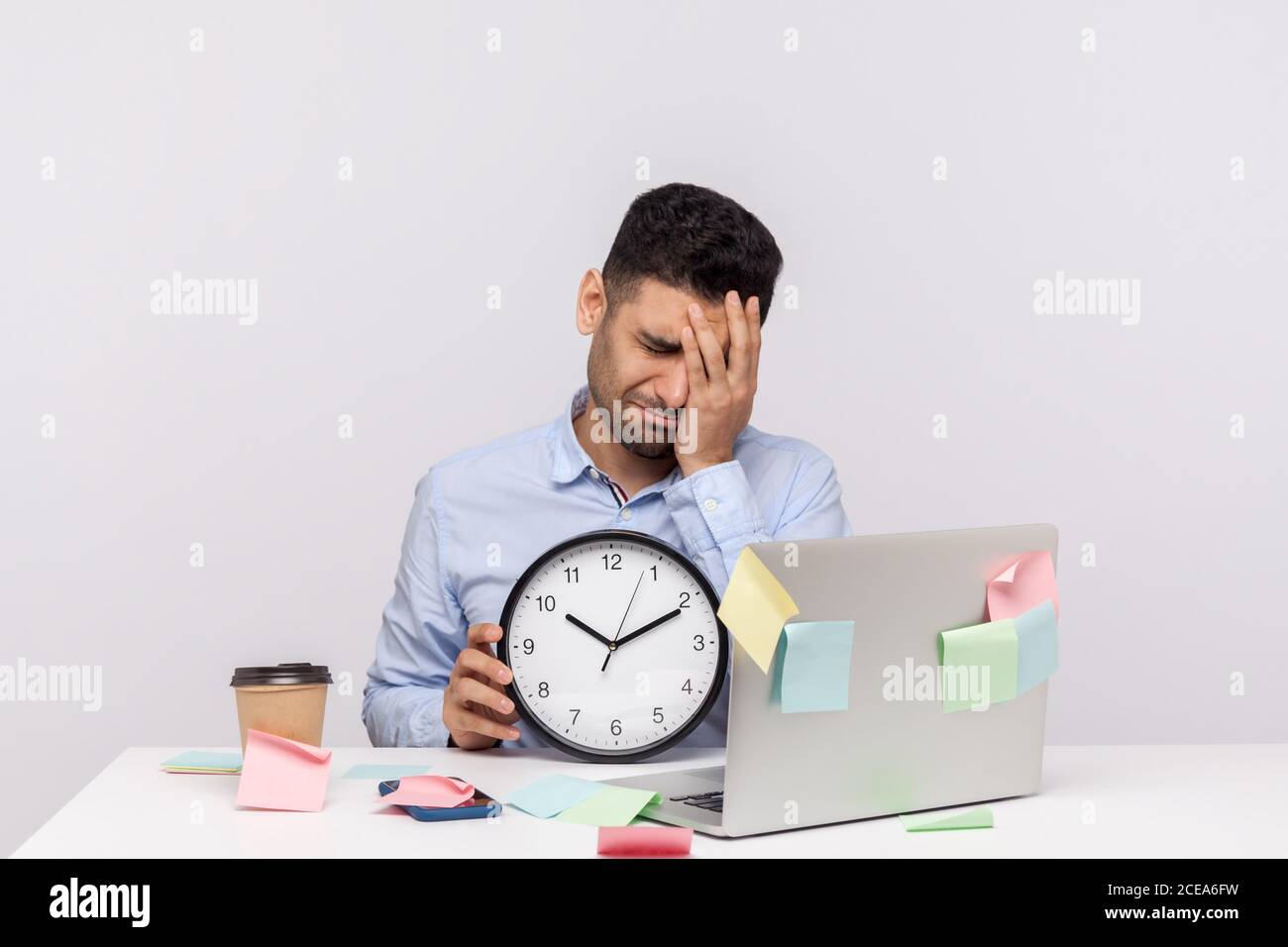 Overtime work. Unhappy sorrowful man employee sitting in office workplace, holding clock and keeping facepalm gesture, expressing regret of missed dea Stock Photo