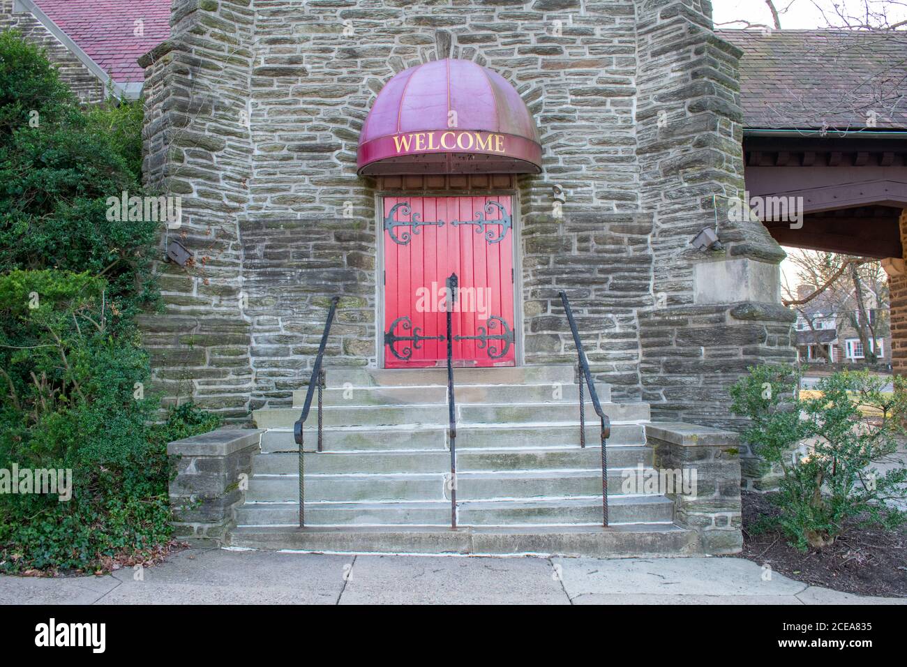 A Bright Red Door in a Cobblestone Wall With a Welcome Sign Above and Steps Leading Up to It and a Bush on the Side Stock Photo