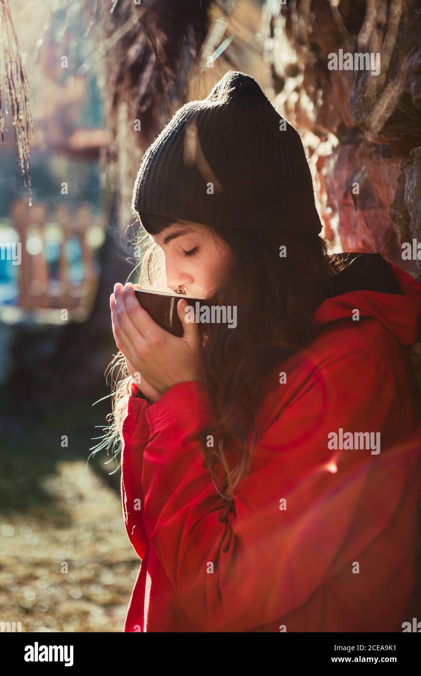 Side view of beautiful young female in stylish hat keeping eyes closed and smelling aromatic hot drink while sitting in countryside on sunny day Stock Photo