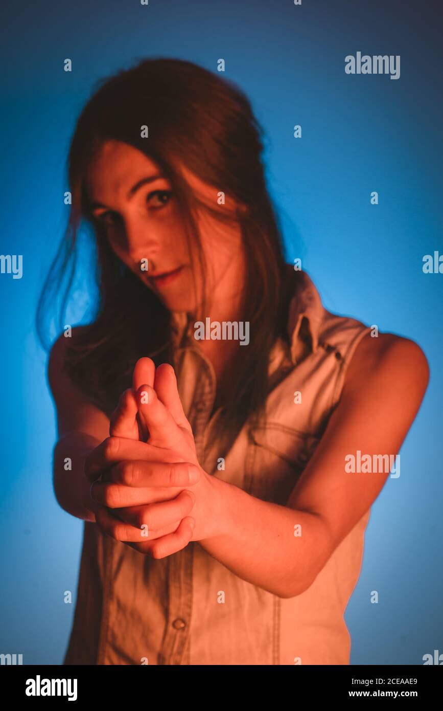 Charming young lady with piercing showing gun gesture and looking at camera in redness on blue background Stock Photo