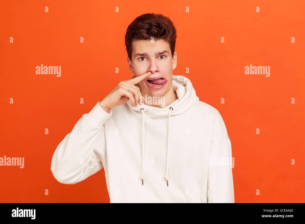 Mischievous teenager in white casual style hoodie putting finger into his nose and showing tongue, fooling around, bad habits, disrespectful behavior. Stock Photo