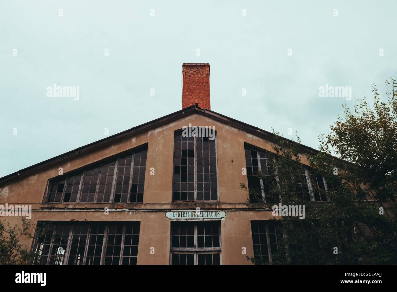 From below red facade of aged broken construction with big windows and chimney near trees and cloudy heaven Stock Photo