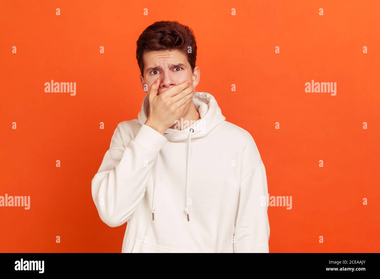Oh my God, unbelievble! Shocked young man with stylish hairdo in casual hoodie closing mouth with hand, keeping secret information. Indoor studio shot Stock Photo