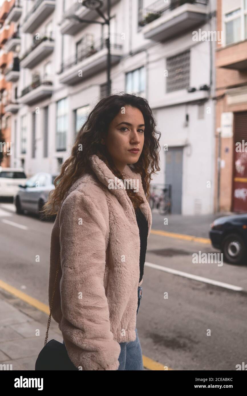 Beautiful young Woman with curly hair looking at camera while standing near modern building on city street Stock Photo