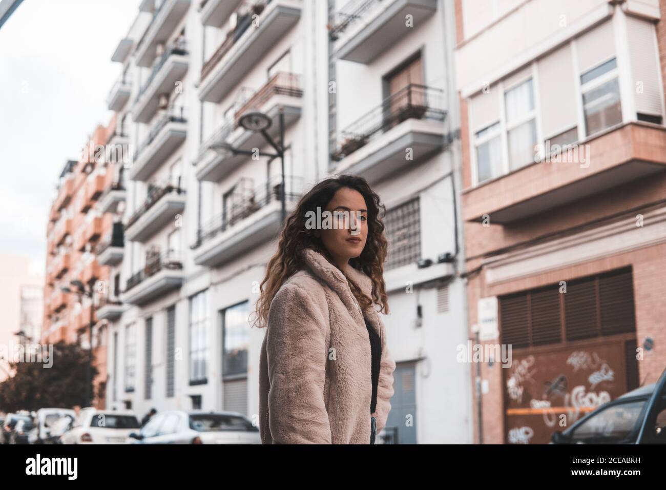Beautiful young Woman with curly hair looking at camera while standing near modern building on city street Stock Photo