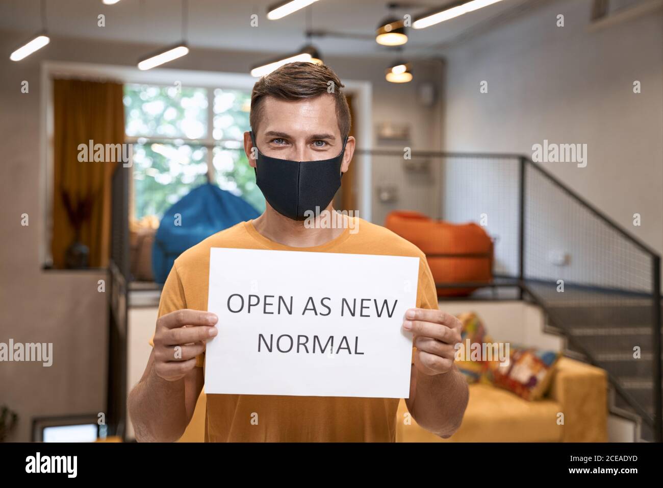 Social distancing at work. Young male office worker in protective face mask showing paper with text OPEN AS NEW NORMAL at camera while standing in the Stock Photo