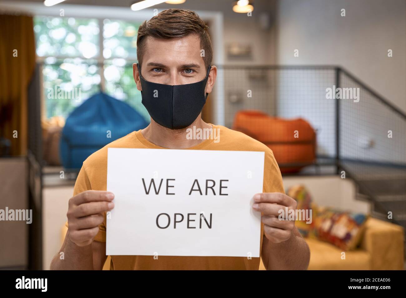 Young male office worker in black protective mask showing paper with text WE ARE OPEN at camera, standing in the modern office Stock Photo