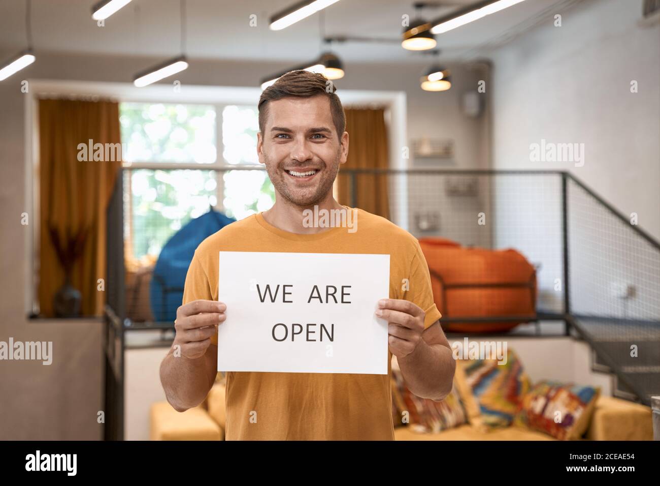 Opening office after lockdown. Young happy man showing paper with text WE ARE OPEN at camera and smiling while standing in the modern coworking space Stock Photo