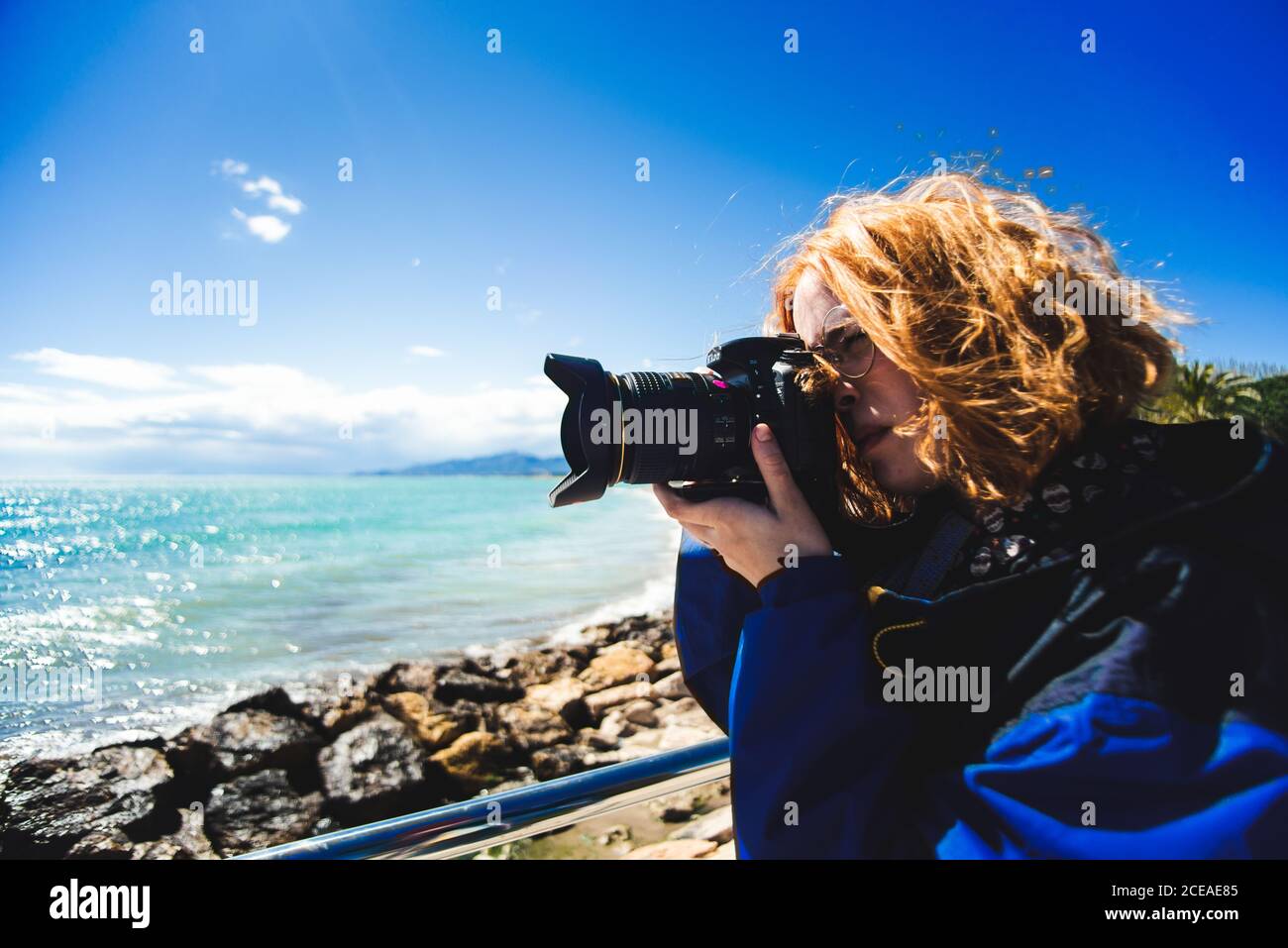 Side view of pretty redhead Woman taking shots of seascape on rocky coast in sunny day. Stock Photo