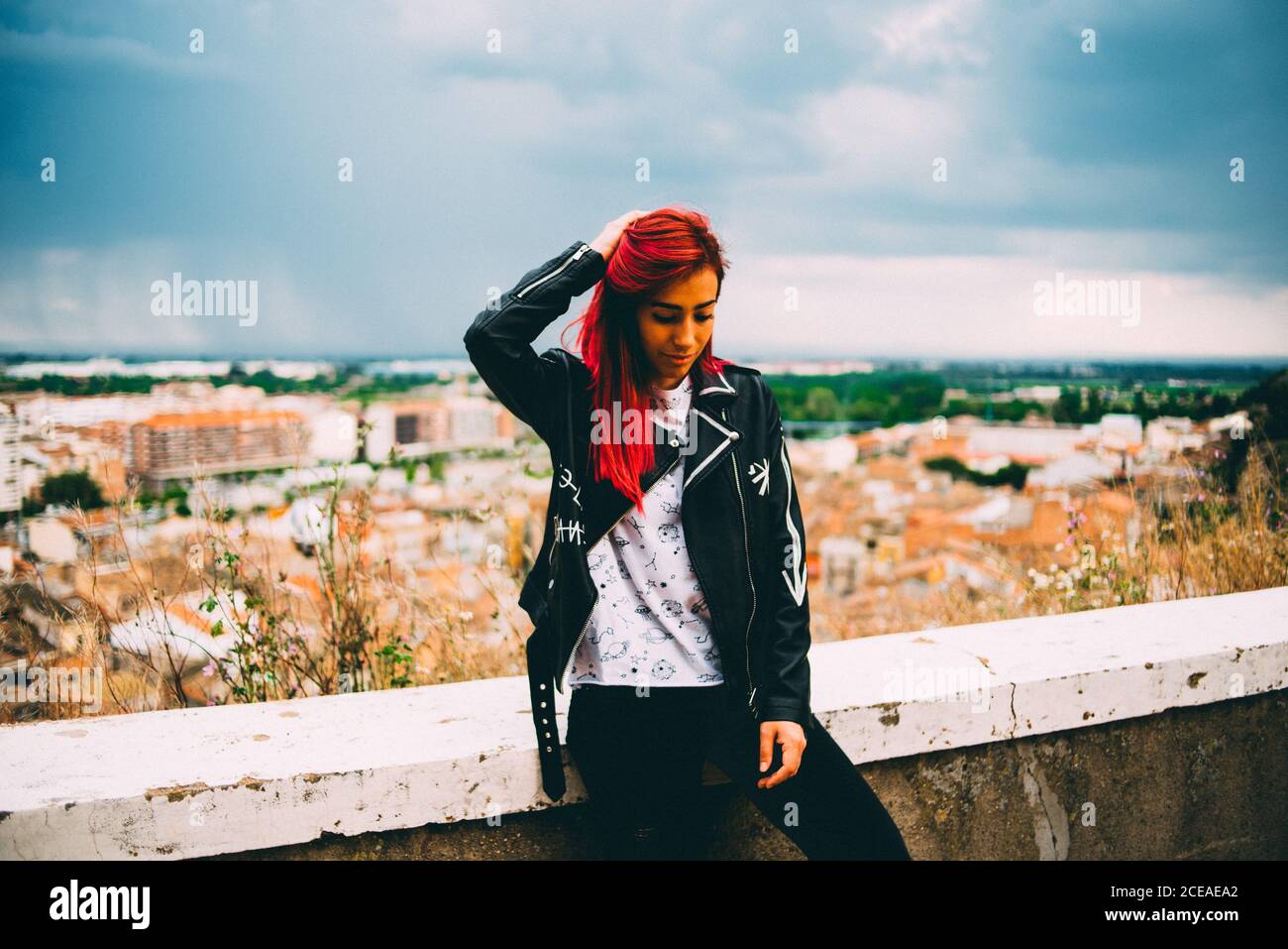Stylish Woman in trendy jacket and with red dyed hair standing on terrace with cityscape in overcast Stock Photo