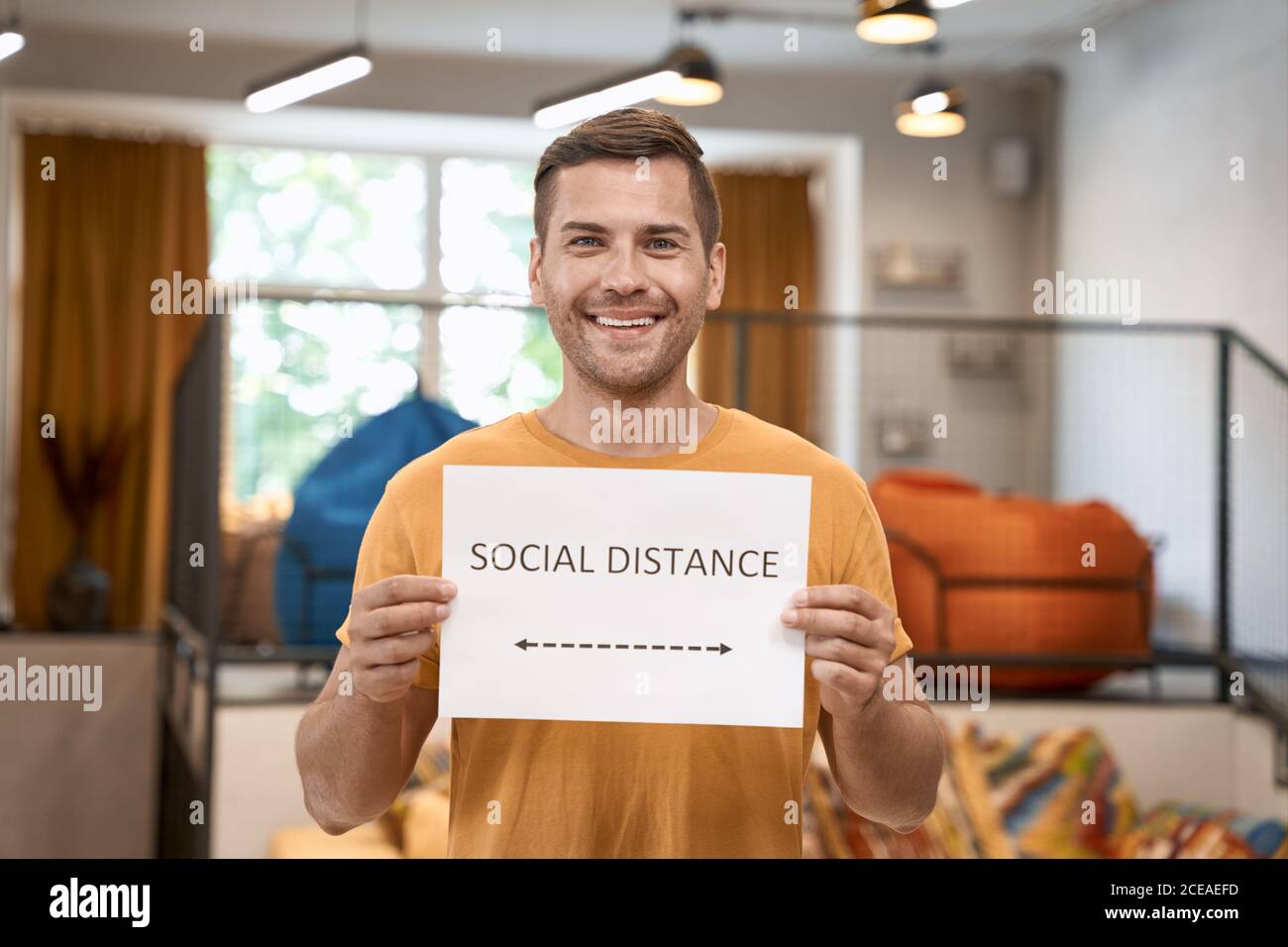 Reopening office safely. Young happy man showing paper with text SOCIAL DISTANCE at camera and smiling, standing in the modern coworking space Stock Photo
