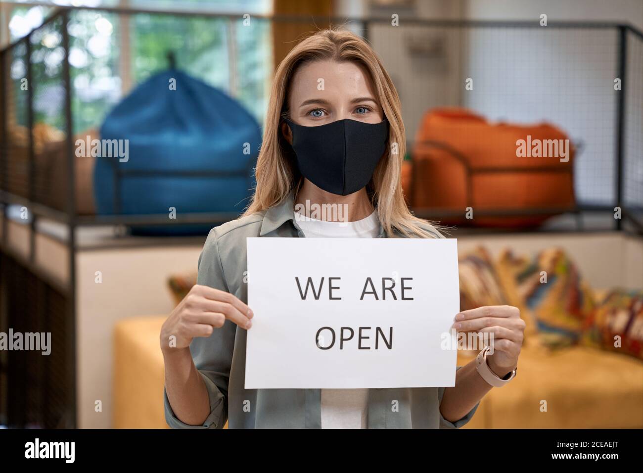 Young caucasian woman, female office worker in protective face mask showing paper with text WE ARE OPEN at camera while standing in the modern office Stock Photo