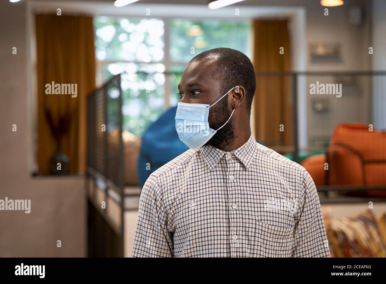 Working safely during coronavirus. Portrait of young afro american man, male office worker wearing protective face mask and looking aside, standing in Stock Photo