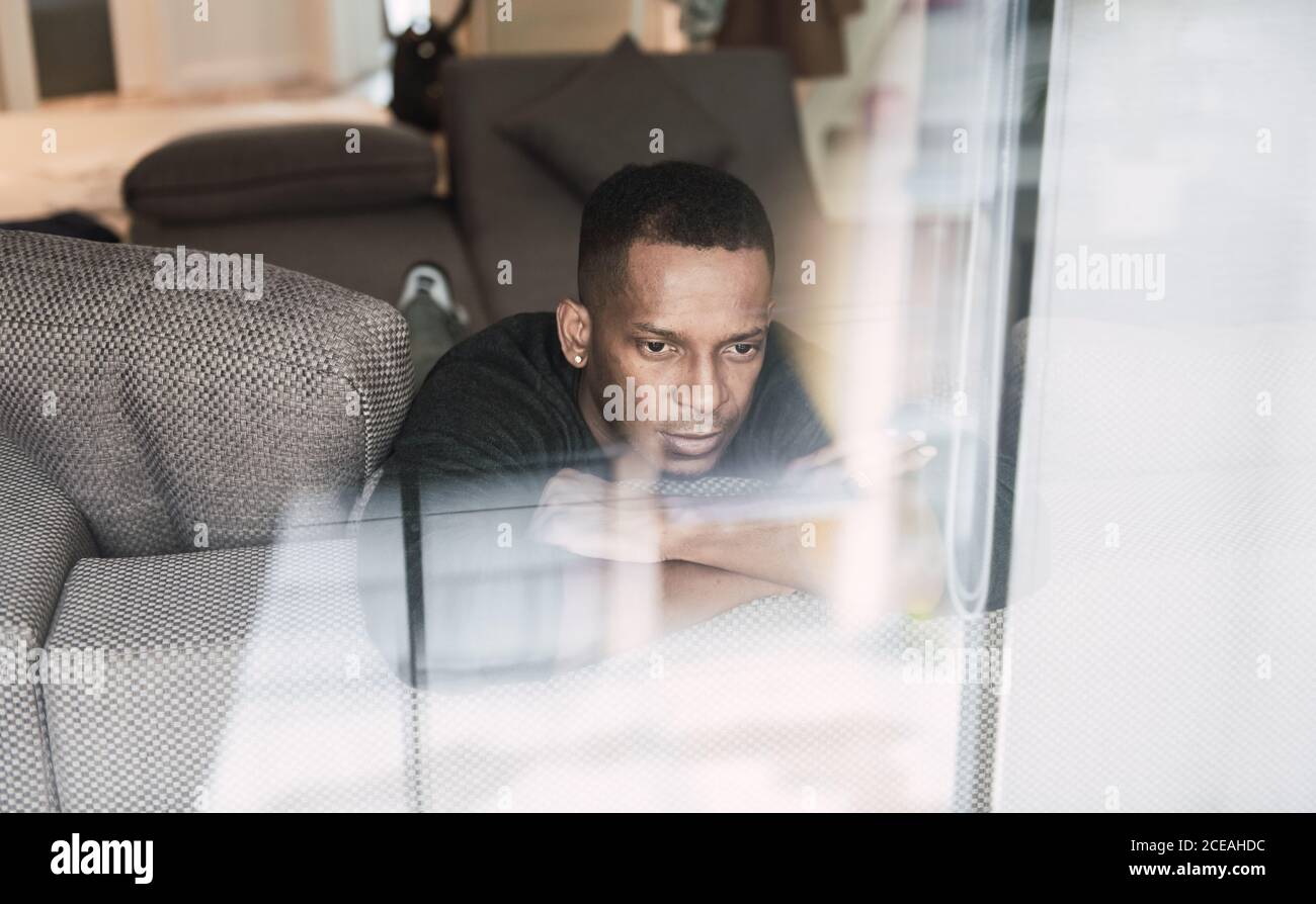 Dreaming adult black man leaning on hands while sitting on couch and looking away in shiny window Stock Photo