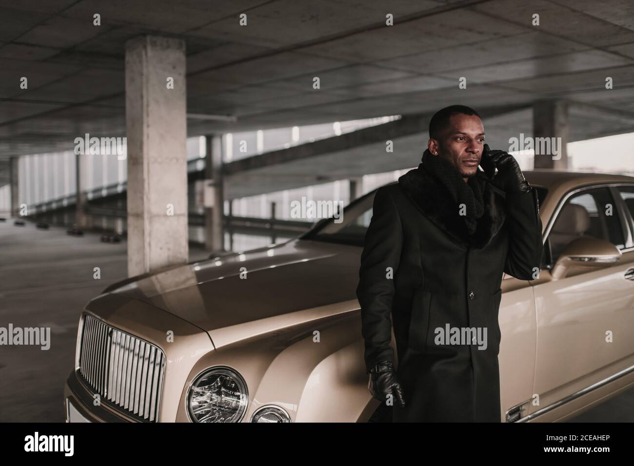 Adult African American male in stylish coat standing near luxury vehicle on parking lot and speaking on smartphone Stock Photo