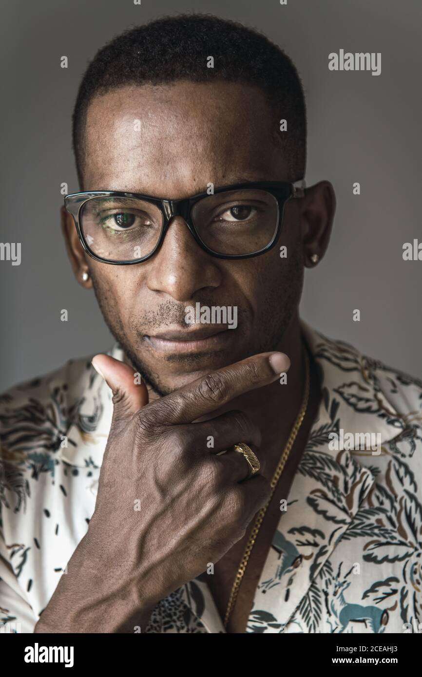 Head shot of serious adult African American man in golden accessories and glasses touching chin Stock Photo