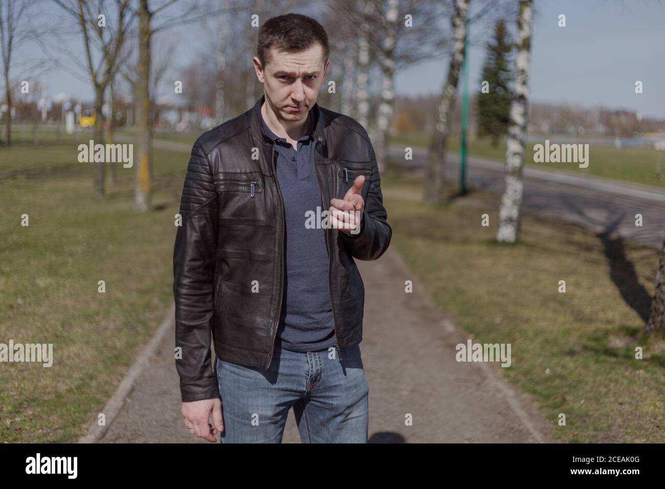 Young adult man in a black leather jacket and jeans walks along the path on a sunny day Stock Photo