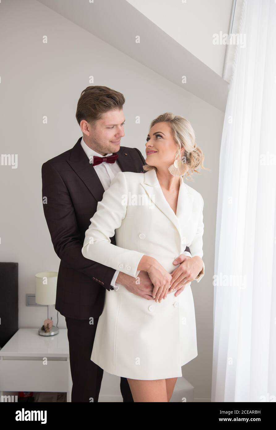 Young handsome man in black costume and beautiful Woman in white jacket standing in room near window and hugging Stock Photo