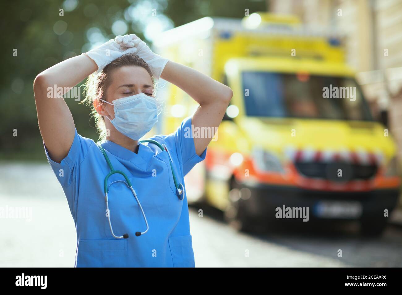 coronavirus pandemic. tired modern paramedic woman in scrubs with stethoscope and medical mask outdoors near ambulance. Stock Photo