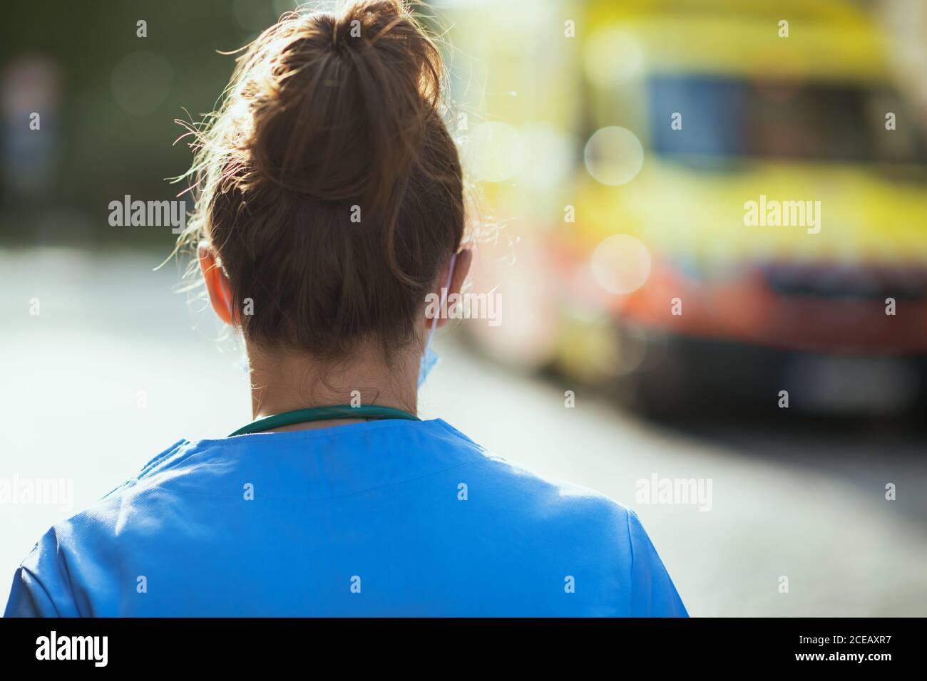 covid-19 pandemic. Seen from behind medical doctor woman in scrubs with medical mask outside near ambulance. Stock Photo